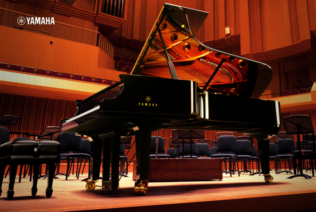 A black Yamaha CFX concert grand piano sits on a wooden stage in a large, empty concert hall with rows of chairs and music stands.