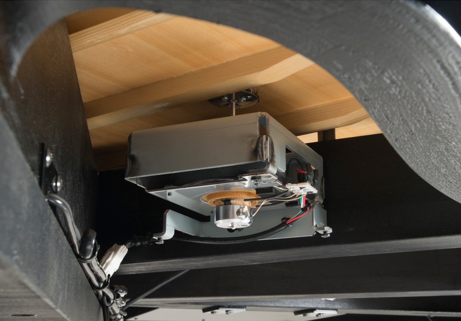 Close-up view of the underside of a Yamaha TransAcoustic piano showing the metal transducer unit mounted to the solid spruce soundboard. The image displays the internal wiring and the mechanism that converts digital sound into acoustic vibrations through the piano's wood.