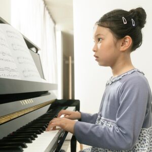 A young girl with her hair in a bun playing a Yamaha B10 upright piano while reading sheet music.
